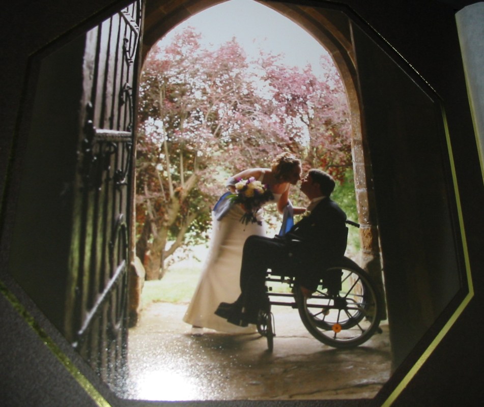 a man in a wheelchair silhouetted in a church doorway, the bride leans forward to kiss him