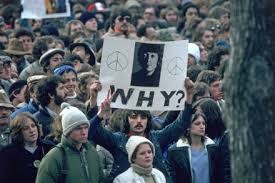 group of people hold a banner. on it is a picture of John Lennon and the word 'why?'