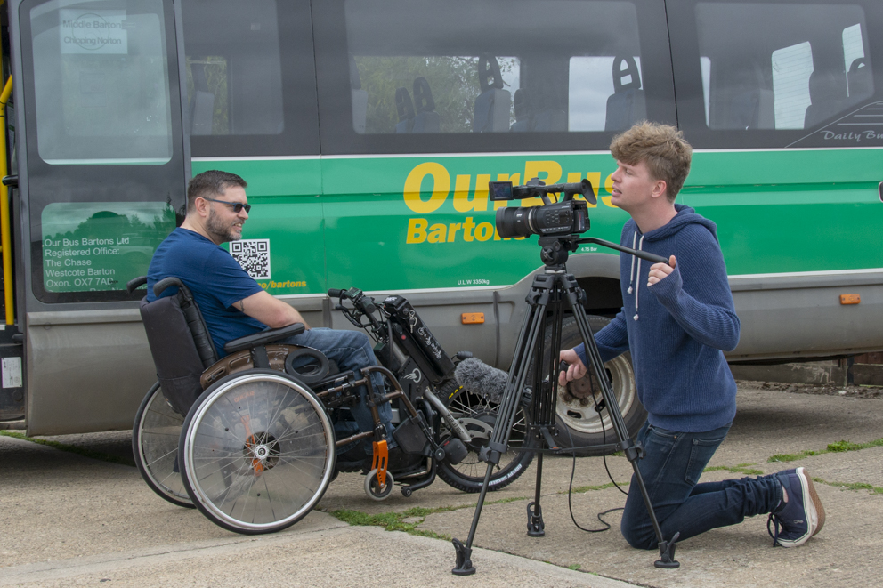 side view of Richard on his trike. a man with a tv camera on a tripod kneels behind itk